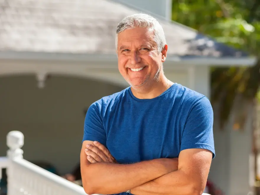 A smiling man with his arms folded, standing outside his home
